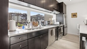 A modern kitchen with dark cabinets, stainless steel appliances, and a large window overlooking a cityscape. - Modern Hideaway with Private Terrace in Downtown (Anchorage)