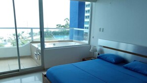 Een slaapkamer met een groot bed, een balkon met uitzicht op palmbomen en gebouwen, en een raam.