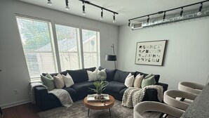 Un salon avec un canapé de couleur sombre, une table basse ronde et une grande fenêtre équipée de stores.