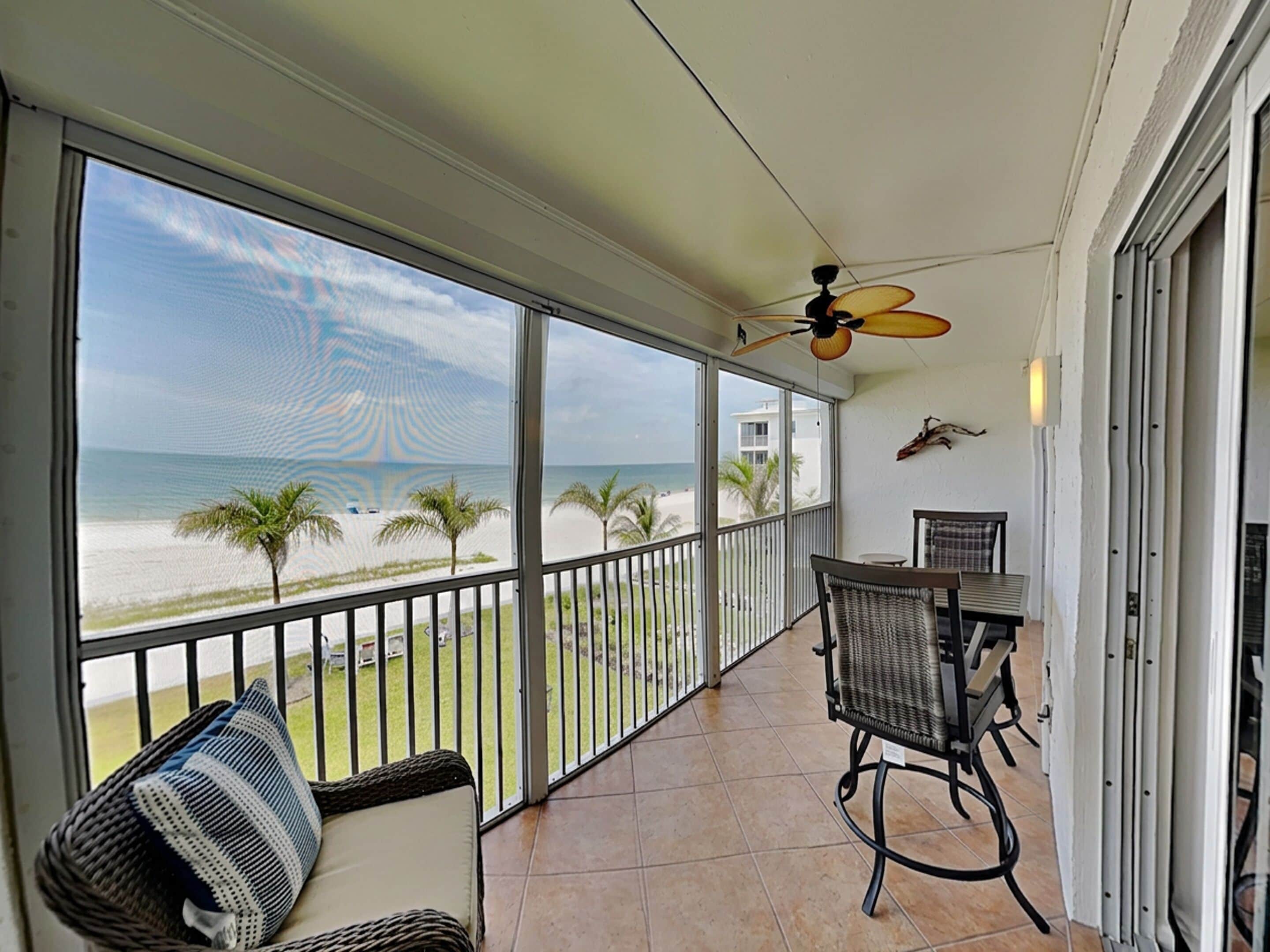 A balcony with a view of the beach, a ceiling fan, and a table with chairs.