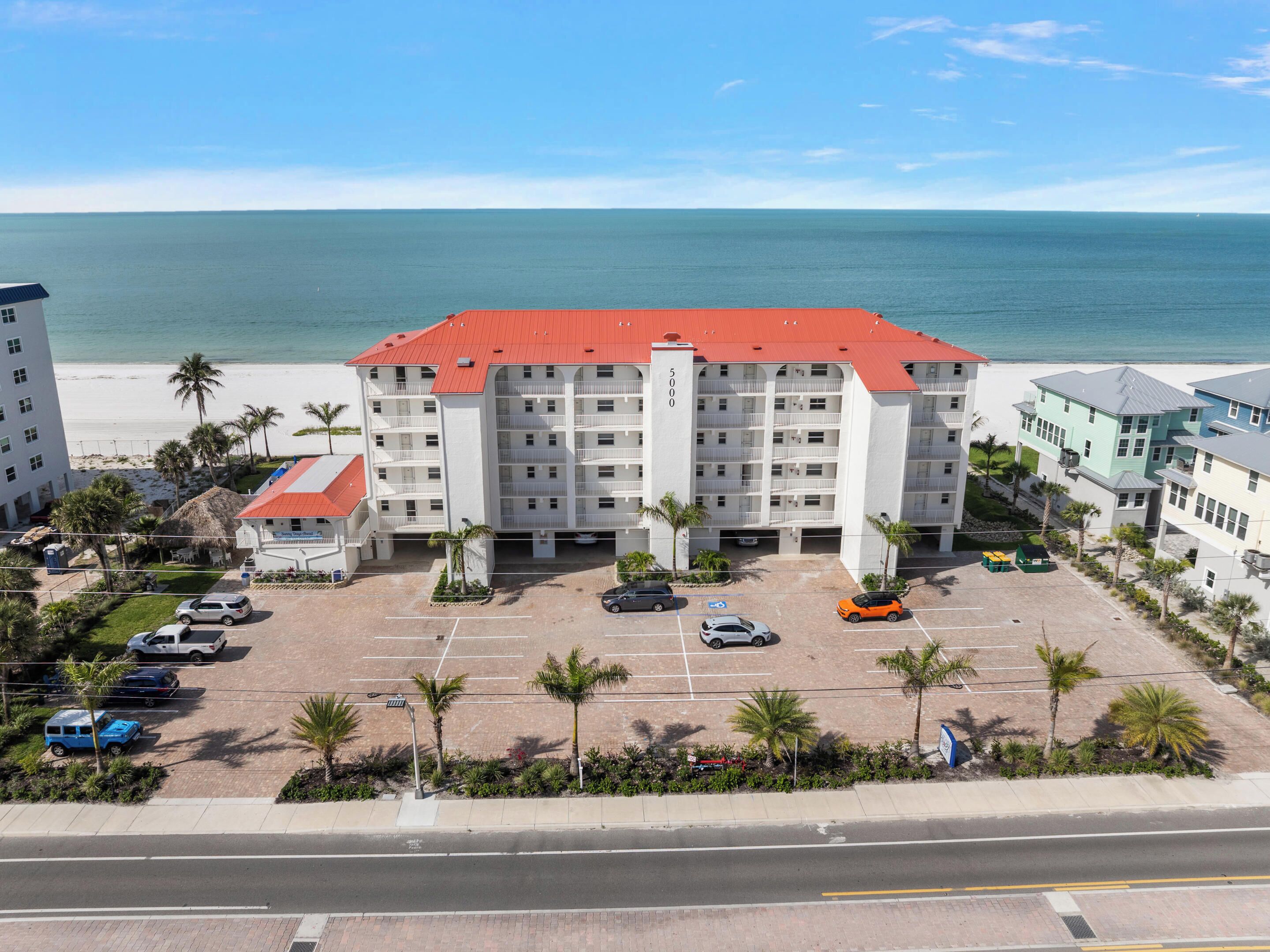 A multi-story hotel with a red roof, a parking lot with several cars, and a view of the beach and ocean.