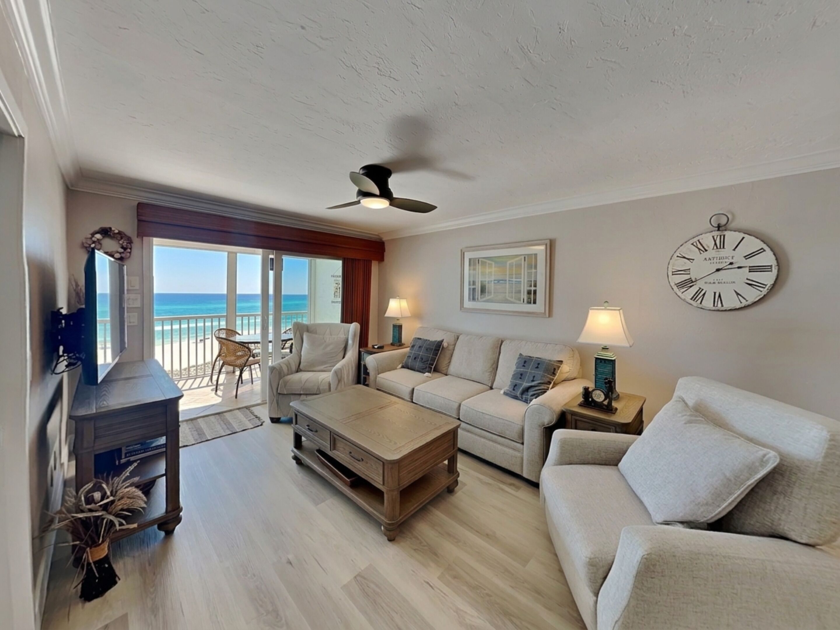 A living room with a sofa, armchairs, a coffee table, a TV, a ceiling fan, a clock, and a view of the ocean.
