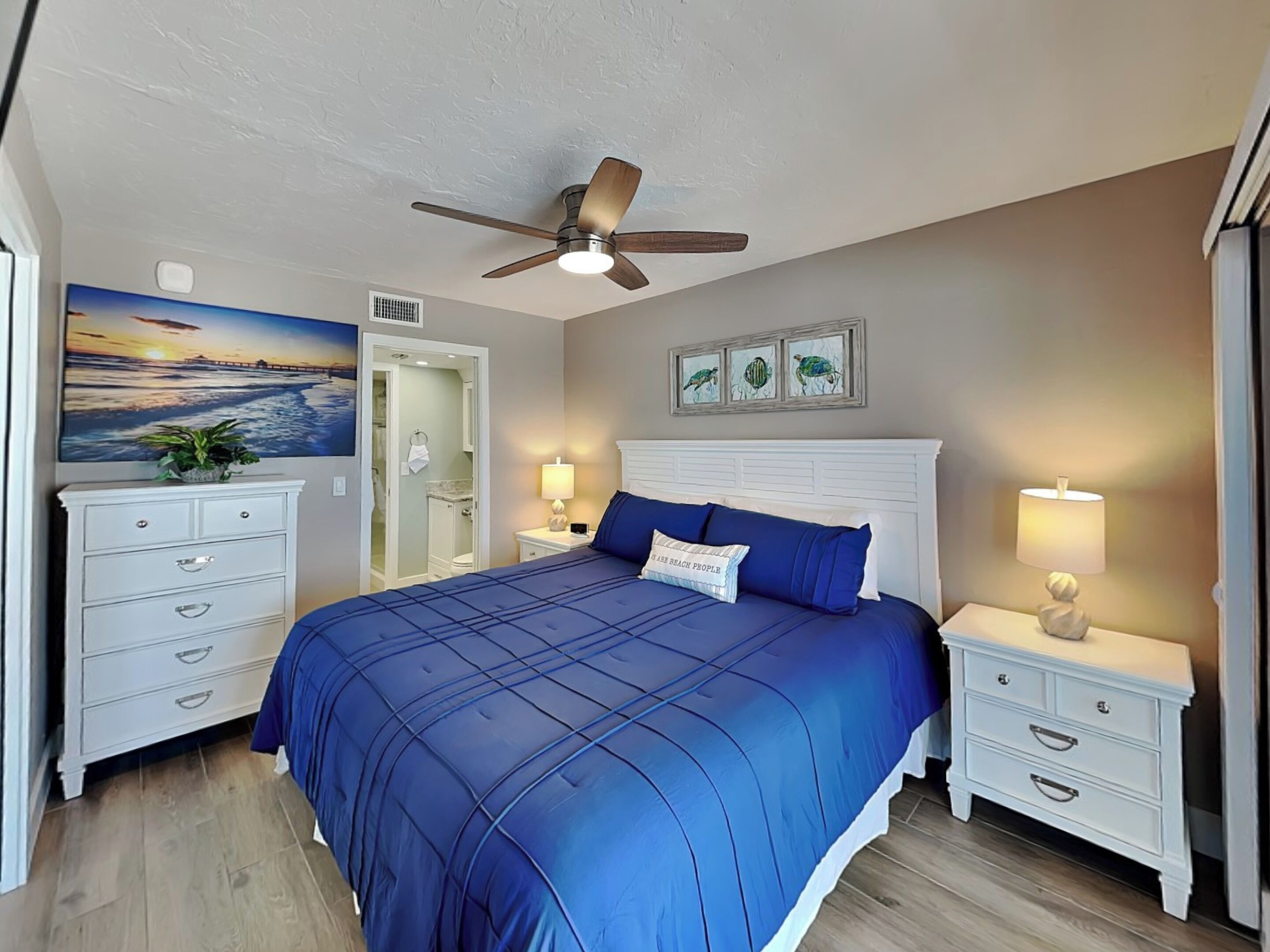 A bedroom with a bed, nightstands, a ceiling fan, a painting, and a door leading to a bathroom.