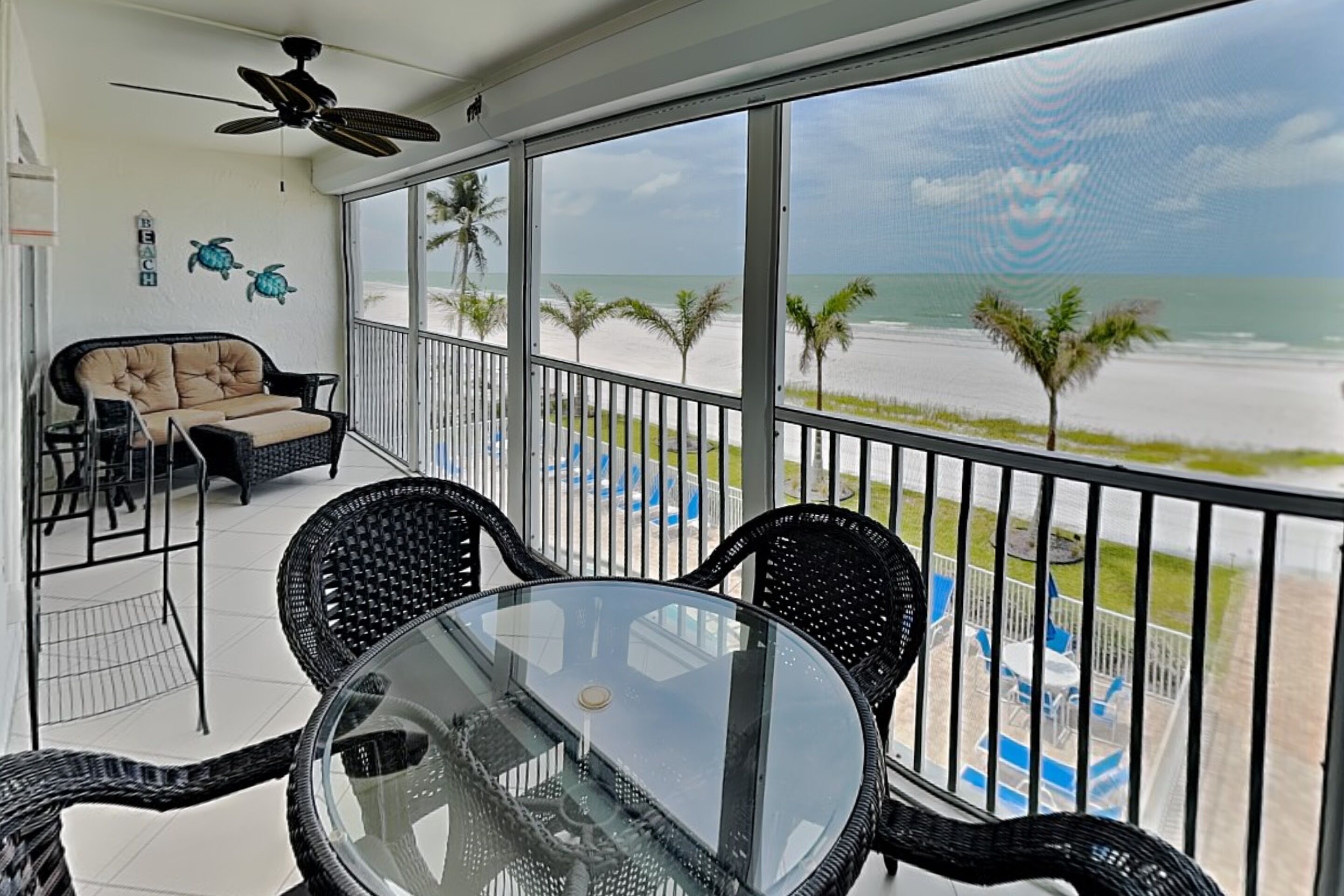 A balcony with a glass table and chairs, a ceiling fan, and a view of the beach.