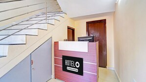 Ein Hotelrezeptionstresen mit einem Schild, das auf betriebsbezogene Leistungen hinweist, eine Treppe mit Metallgeländern und eine Holztür im Hintergrund.