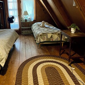 Une chambre mansardée chaleureuse, avec un lit, une petite table ornée d’une plante et un ventilateur.