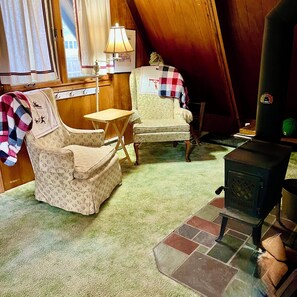 A cozy room with a wooden ceiling, a small table, two chairs, a lamp, and a wood-burning stove.