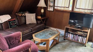 Un salón acogedor en el ático con un sofá de cuero, un sillón mecedor de madera, una mesa de centro de vidrio, una estantería y un televisor.