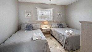 Een kamer met twee bedden, een plafondventilator, een raam met jaloezieën en een nachtkastje met een lamp.