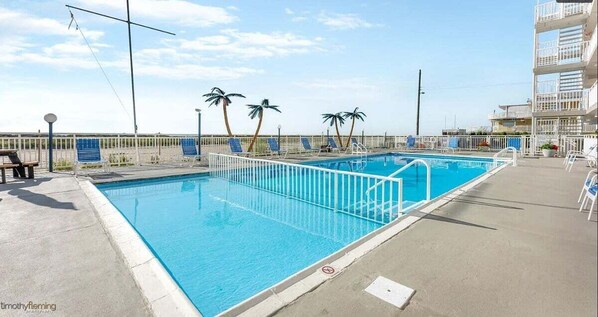 A rectangular pool with a white safety fence, palm trees, and a multi-story building in the background. - 1 Bedroom Beach Condo with a Pool & Balcony (Wildwood Crest)