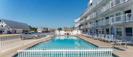 A swimming pool with a white fence, lounge chairs, and a building with balconies.