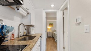 Una cocina con encimera de granito, pileta de acero inoxidable y estufa. Hay un pasillo que conduce a un dormitorio con cama y mesita de noche.