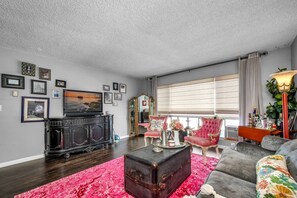 Ein Wohnzimmer mit Fernseher, Schrank, rotem Sessel, Sofa und einem großen Fenster mit Jalousien.