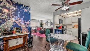 Una habitación con piso de madera, un ventilador de techo, una mesa de comedor con sillas, una zona de cocina y una pared con un mural floral.
