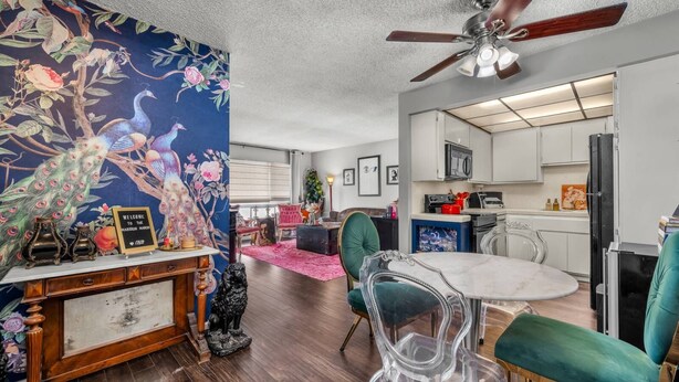 A room with a wooden floor, a ceiling fan, a dining table with chairs, a kitchen area, and a wall with a floral mural.