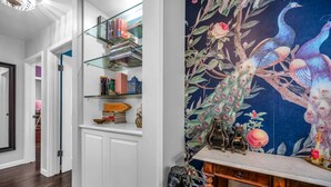 A hallway with a glass shelf displaying books, a decorative peacock mural, and a fireplace with a rustic mantel.
