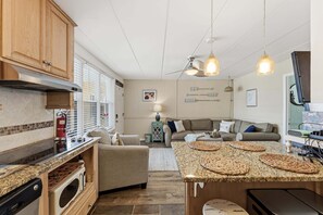 Una cocina compacta con electrodomésticos integrados, un comedor con una isla grande y una sala de estar con sofá y televisión.