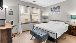 Ein Schlafzimmer mit Bett, Nachttischen, einem Schreibtisch, einer Bank, einem Fenster mit Jalousien und einem Bild an der Wand.