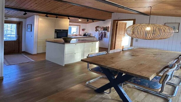 Una cocina amplia con armarios de madera y una isla central, un comedor con una gran mesa de madera y sillas de metal, y una lámpara colgante.