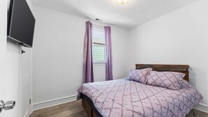 Une chambre à coucher avec un lit, un téléviseur fixé au mur, une fenêtre avec des rideaux et un plancher en bois.