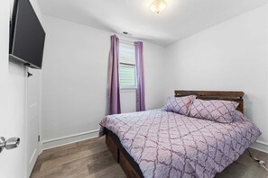 A bedroom with a bed, a TV mounted on the wall, a window with curtains, and a wooden floor.