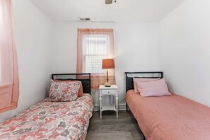A small, single-room bedroom with two beds, a bedside table with a lamp, a window with sheer curtains, and a ceiling vent.