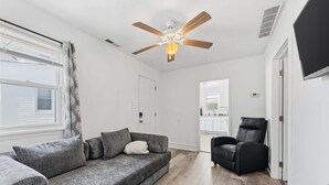Un salon avec un canapé gris, un fauteuil noir, un ventilateur de plafond et une vue sur la cuisine.
