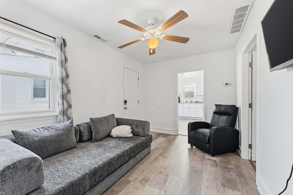 A living room with a grey sofa, a black chair, a ceiling fan, and a view into the kitchen.