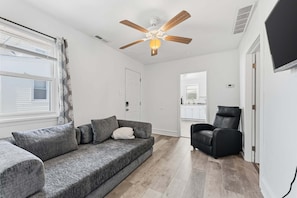 A living room with a grey sofa, a black chair, a ceiling fan, and a view into the kitchen.