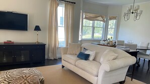 Una sala de estar con sofá, televisión, lámpara, zona de comedor con mesa y sillas, y ventanas grandes con cortinas.