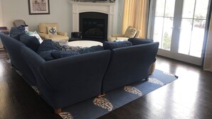 Un salón con un sofá azul oscuro, una chimenea, un televisor y un amplio ventanal con cortinas.