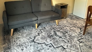 Un sofá gris con patas de madera, una alfombra estampada, una pequeña mesita de madera y un electrodoméstico blanco en una habitación con paredes lisas.
