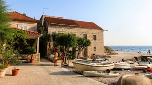 A stone building with a red-tiled roof, a paved area with potted plants, and boats on a beach. - Apartments Trojanović (Orebic)