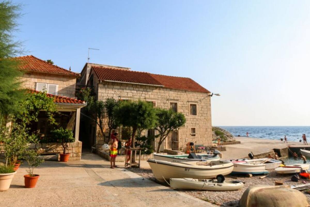 A stone building with a red-tiled roof, a paved area with potted plants, and boats on a beach.