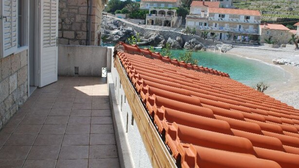 A balcony with terracotta tiles, a stone building, and a view of a beach and hillside buildings. - Apartments Trojanović (Orebic)