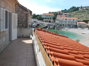 A balcony with terracotta tiles, a stone building, and a view of a beach and hillside buildings. - Apartments Trojanović (Orebic)