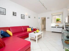 A red sofa with yellow and blue cushions, a white coffee table, and a kitchenette with a window. - Beach House Legosi (Dubrovnik)