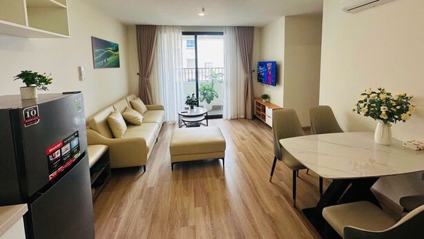 A modern living room with a sofa, ottoman, dining table, and chairs. There is a refrigerator, a TV, and a plant.