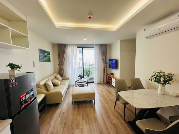 A modern living room with a sofa, ottoman, dining table, and chairs. There is a refrigerator, a TV, and a plant.