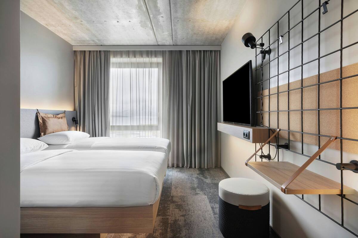Habitación de hotel con dos camas, un televisor montado en una pared de paneles y un pequeño estante de madera, además de una ventana con cortinas.