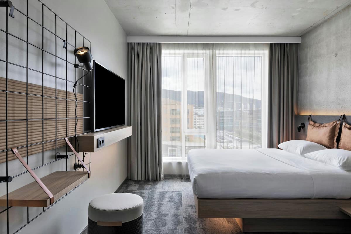 Una habitación de hotel moderna con una cama grande, un televisor montado, una ventana con cortinas y vistas a la ciudad.