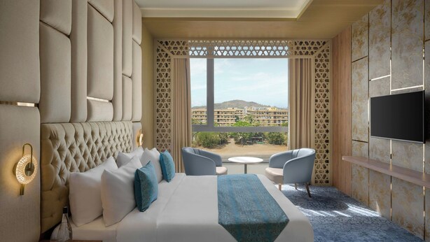 Habitación de hotel con una cama grande, televisión, balcón con vistas a edificios y un papel tapiz estampado.