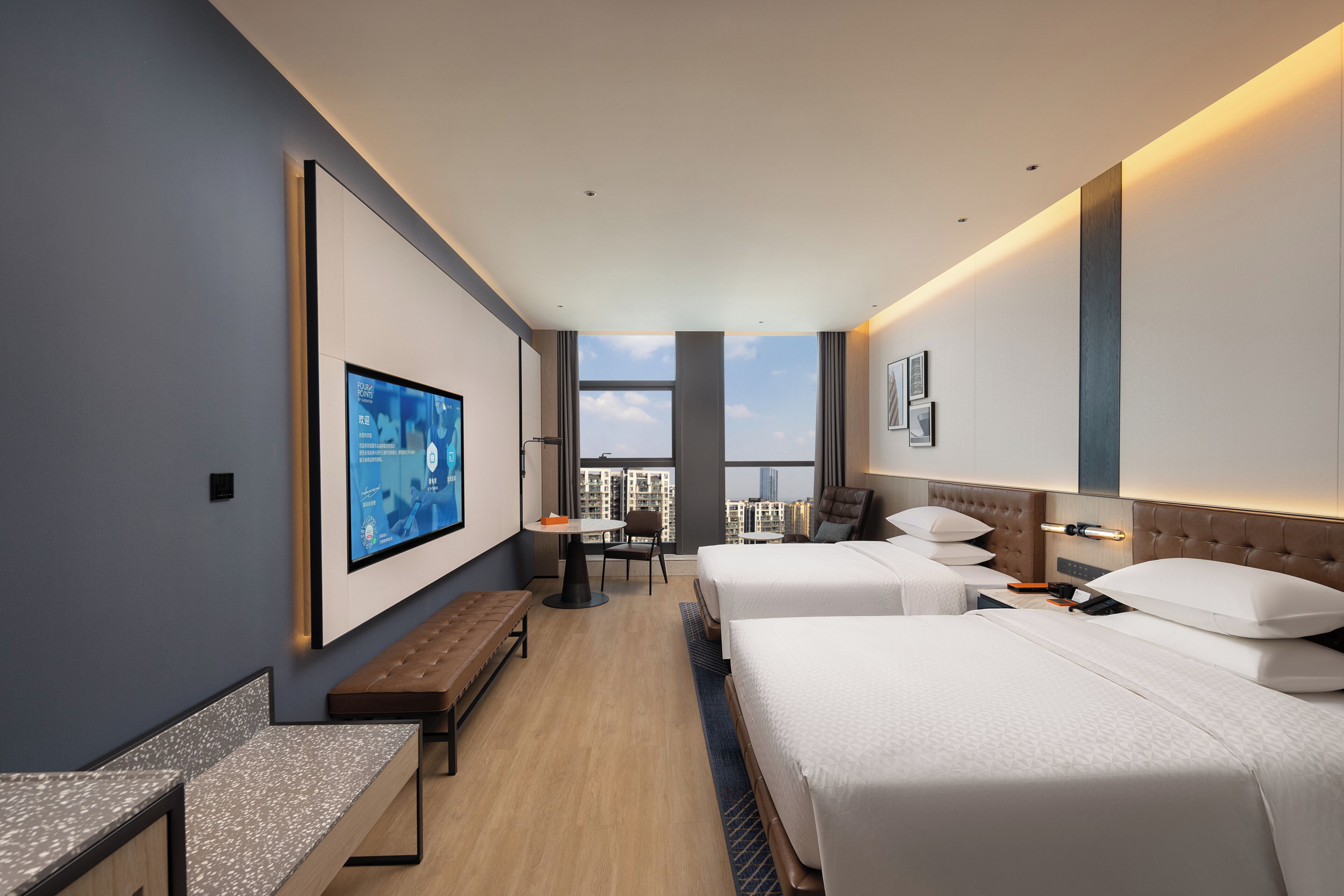 A modern hotel room with two beds, a desk, a bench, a TV, and a view of the cityscape.