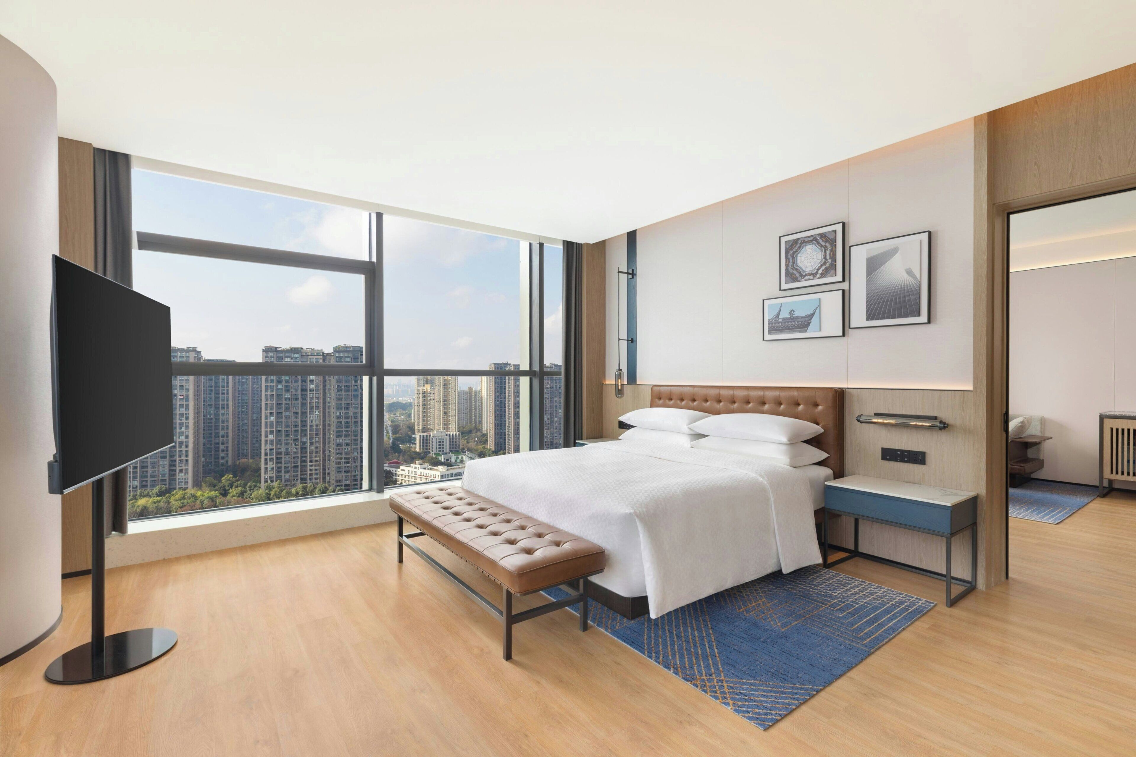 A modern hotel room with a large window, a bed, a bench, a TV, and a small table.