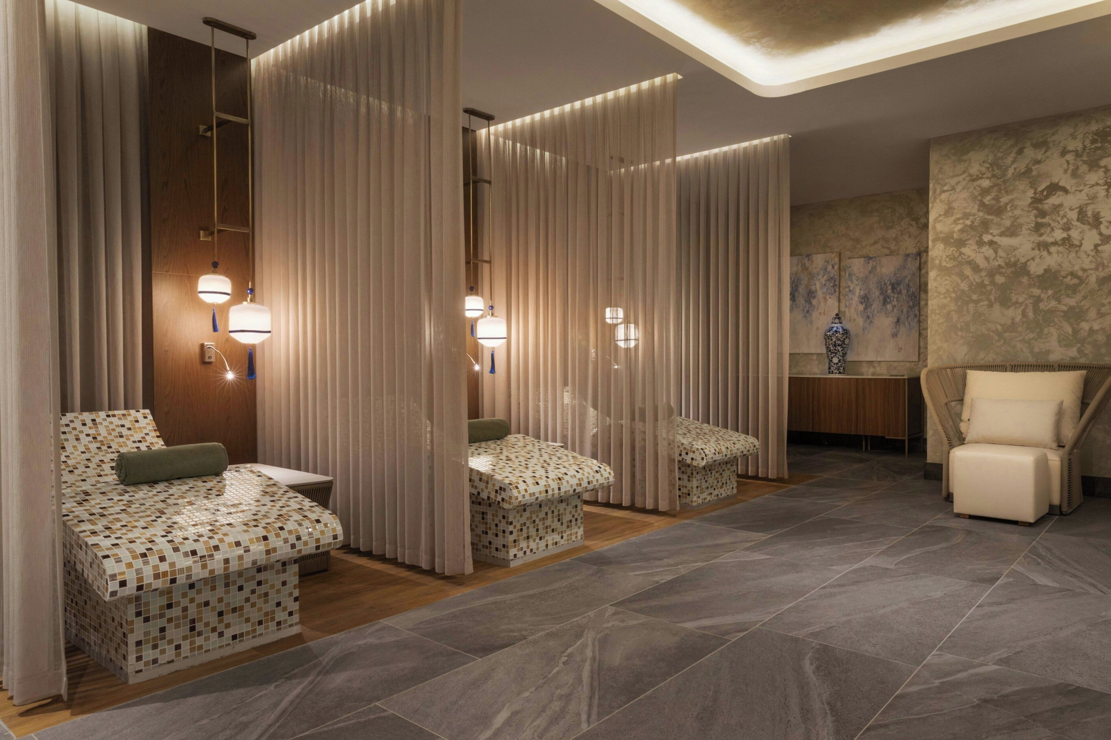 A modern spa room with a bed, two cushioned seating areas, a massage table, and a chair.
