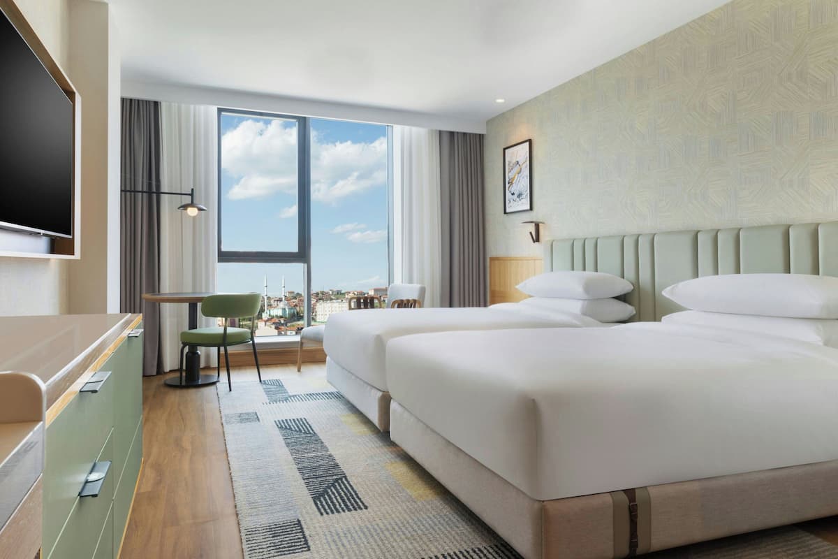 Une chambre d’hôtel avec deux lits, un bureau, une chaise, une télévision et une grande fenêtre donnant sur la ville.