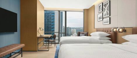 Une chambre d’hôtel avec deux lits, un bureau, une chaise, un banc et une grande fenêtre donnant sur la ville.