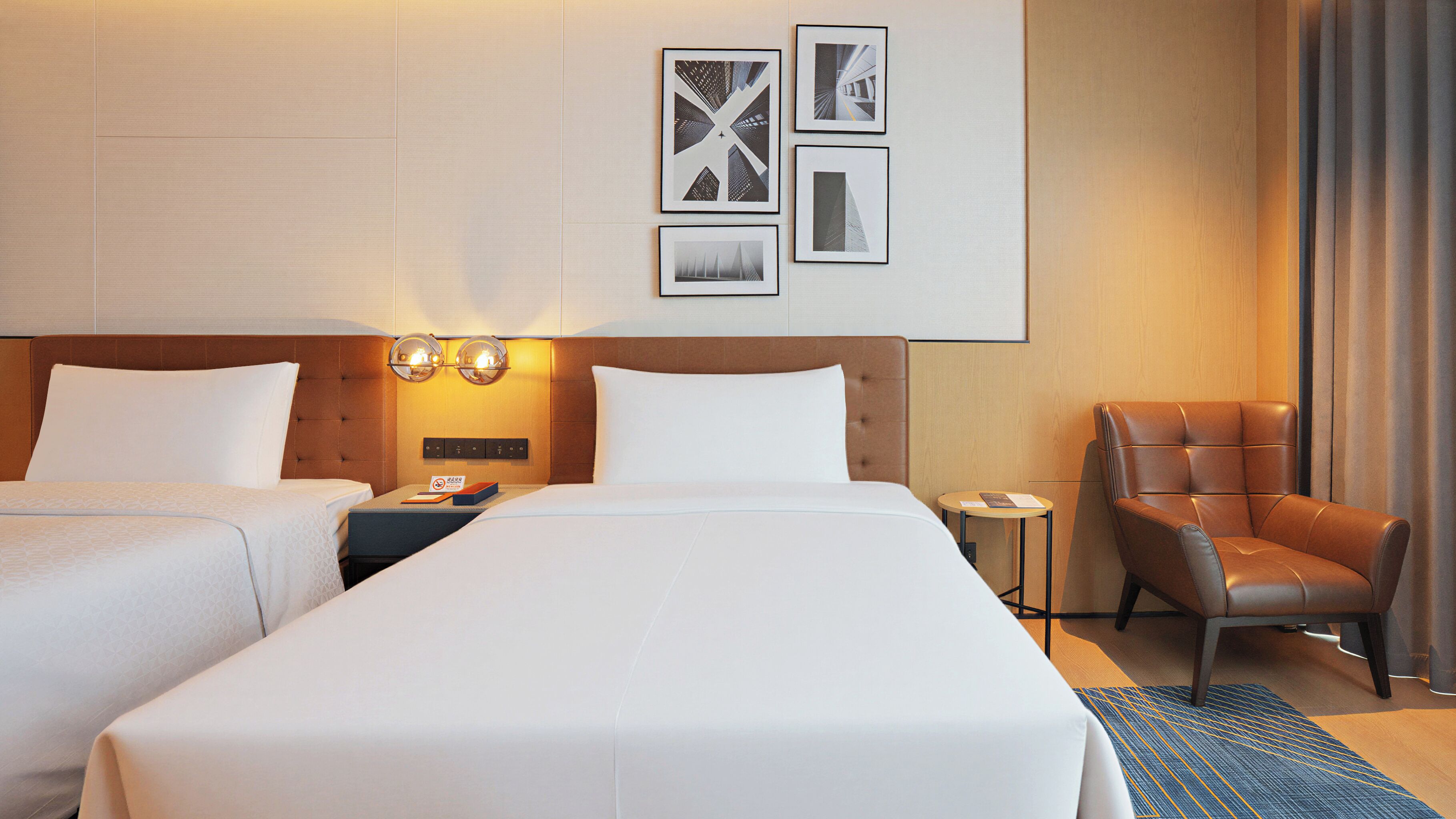 Une chambre d’hôtel avec deux lits, un fauteuil en cuir marron, une petite table et des tableaux encadrés au mur.