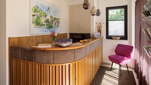 Un mostrador de recepción con detalles de madera, una silla rosa, un cuadro de un edificio y una ventana con cortinas.