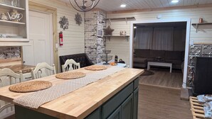 A kitchen with a wooden island, a dining table, and chairs. There is a fireplace, a wall-mounted shelf with decorative items, and a hanging flower.